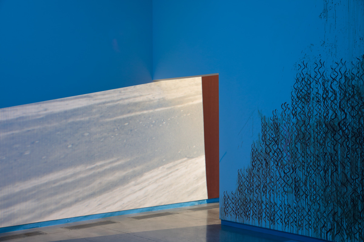 a gallery space with bright blue walls and intricate dark waves that swirl up like flames on the right wall, there is an entry way and the bright projection of a film in the adjacent room leaks through.