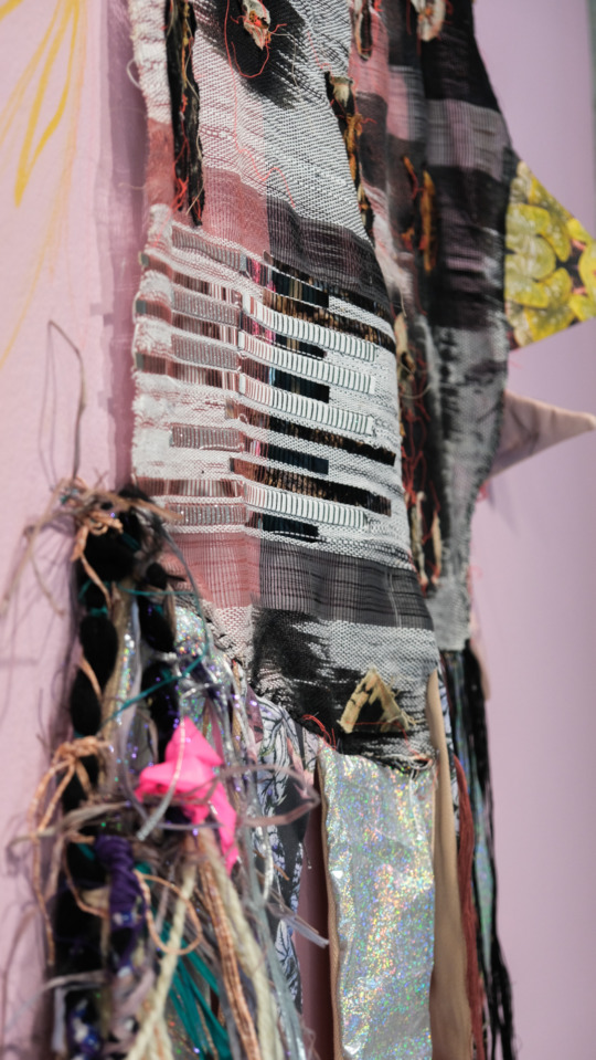 a chaos of fabric, woven into a large textile with holographic thread, screen-printed fabric and black material, a detail shot of the work on a light pink exhibition wall