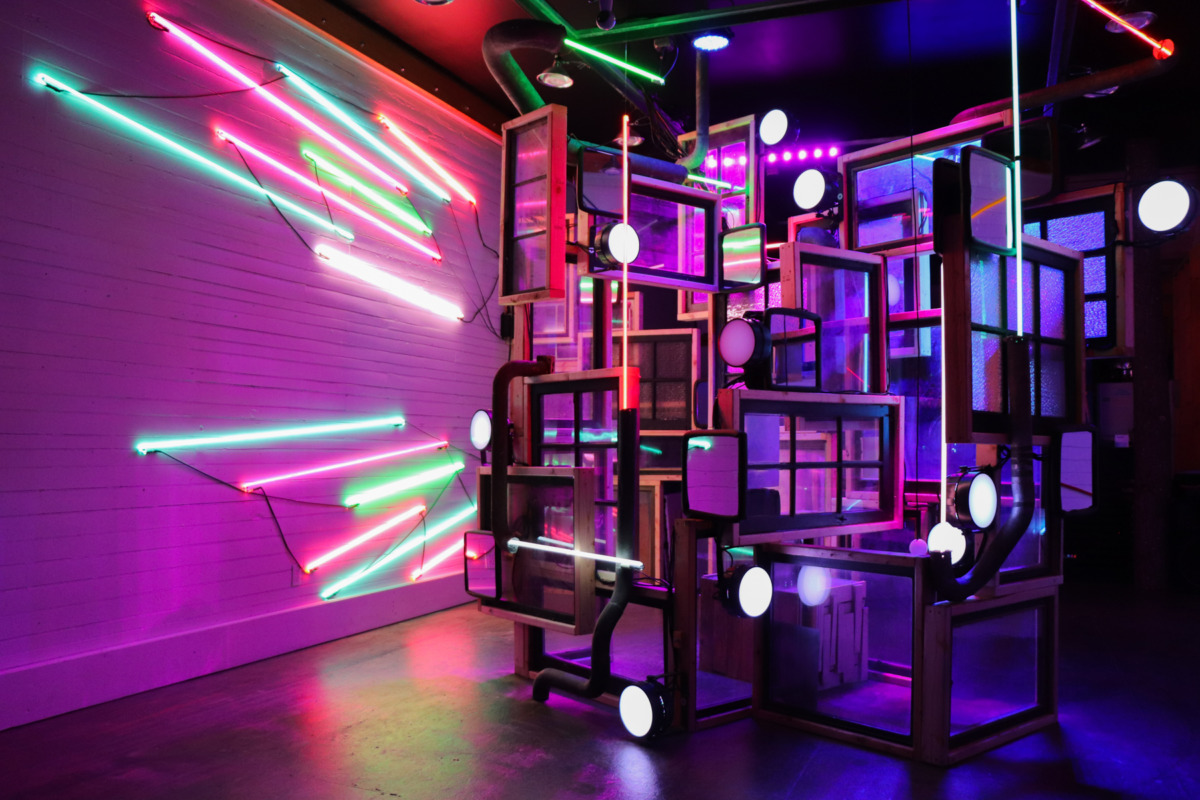 a gallery space with neon LED lights on the walls, and a large collage sculpture made up of window panes. the black flooring and white wall are bathed in bright majenta light with streaks of lime green