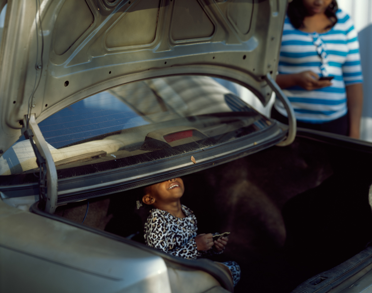 image of a small Black child inside the trunk of a sedan, the trunk lid is open and the small child grins up at the camera, too short for us to see their eyes