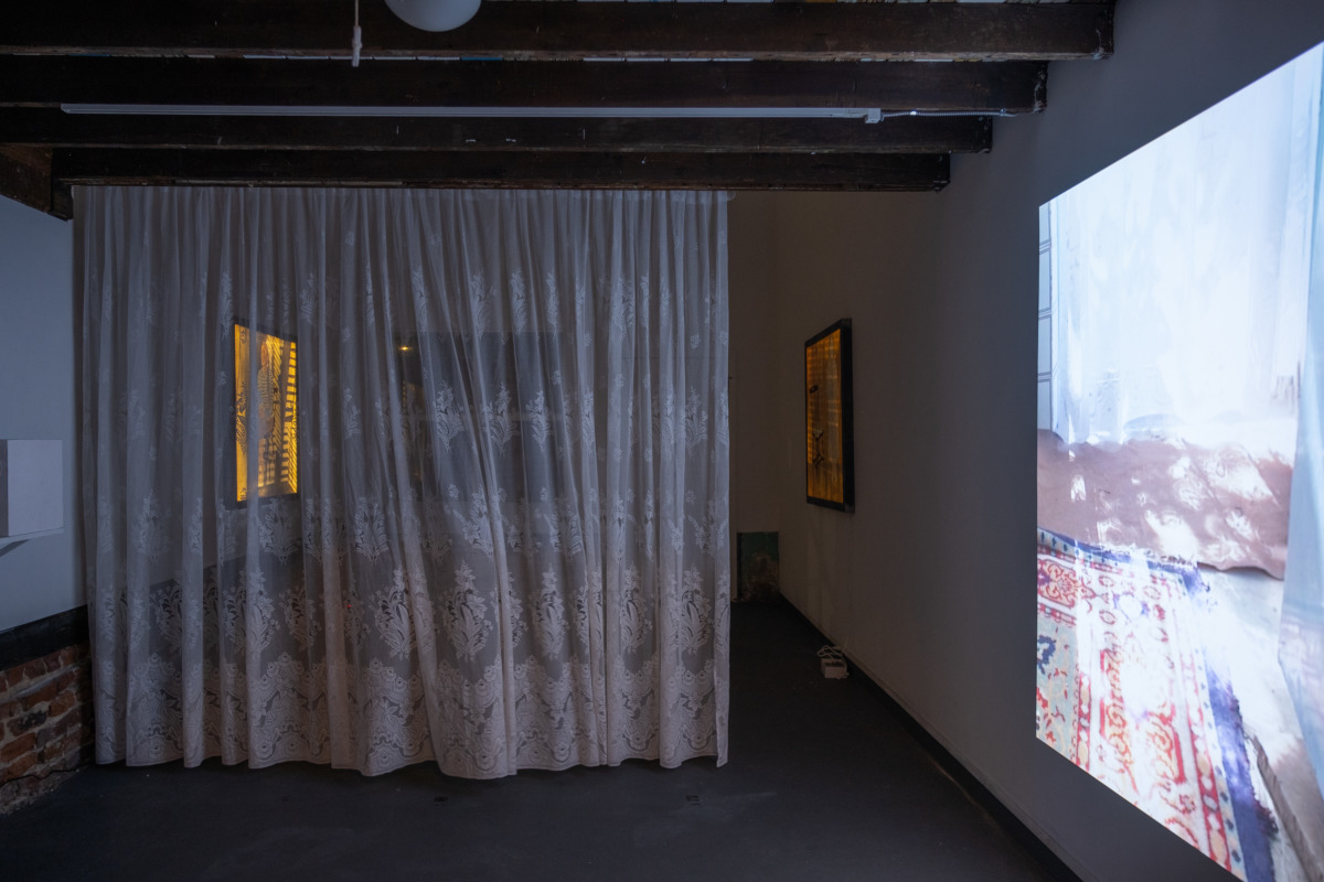 photo of a dim gallery space filled with a white lace curtain, a projected video, and two golden posters on white walls with exposed brick