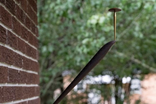 a photograph of a flagpole on the side of a brick house, holding a large nail. full green trees are behind the flagpole