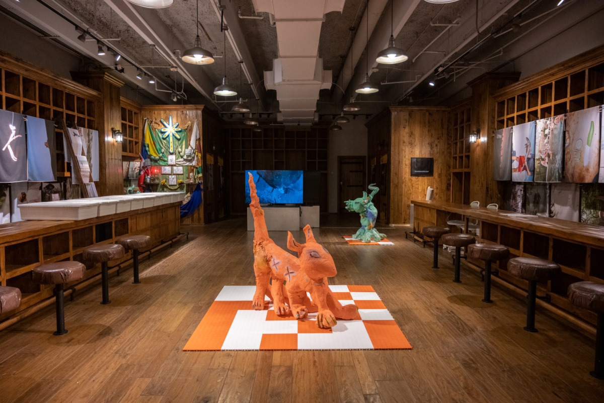 a wood paneled room with brown bar stools. in the middle of the room an orange dog like animal with six legs stands on an orange and white checkerboard. in the back of the room a similar figure in green stands on a checkerboard.
