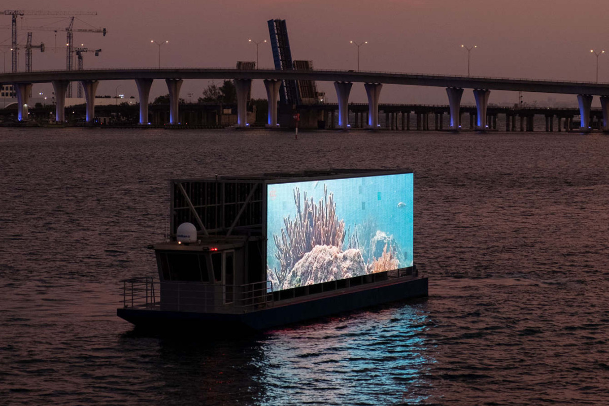 Biscayne Bay at dusk,  with a bridge stretching across the horizon. in the center of the picture is a boat with a video screen broadcasting images of coral. 