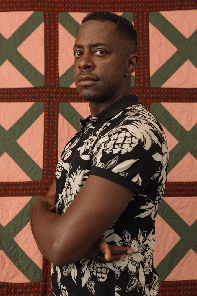 a photo portrait of a Black man wearing a black and white tropical print button down. he is standing against a pink quilt with dark olive green x's and red diamond print grids