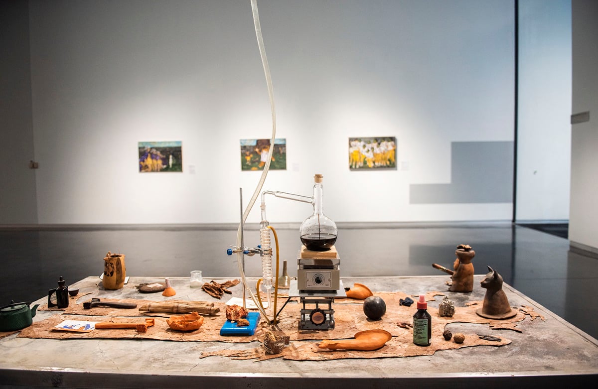 installation view of a white gallery space, in the foreground is a steel table with a large tobacco leaf silhouette of a human, atop the silhouette are a number of objects including herbs, tools, figurines, and a large distillation system