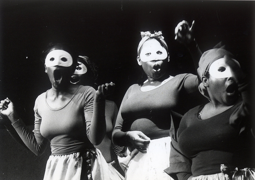black and white photo of Black women in white masks, black shirts and white skirts with their mouths open in a yell. 