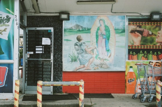 exterior of a bright red brick building with colorful Taki's, Fanta, and Coca-Cola advertisements. In the center of the wall is a painting of Our Lady of Guadalupe appearing to a Mexican man named Juan Diego in the miracle of roses.