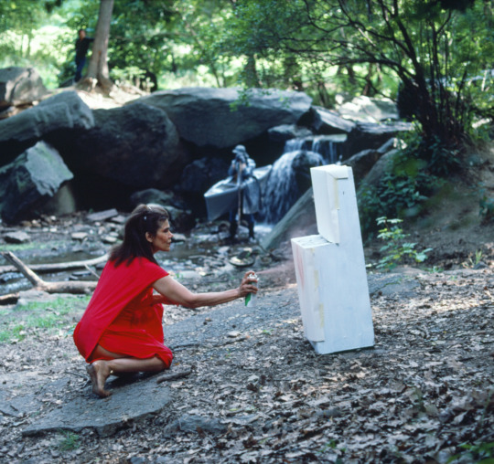 an older Black woman wears a bright red dress in a forest. she kneels down next to a white wooden object and begins to spray paint it red
