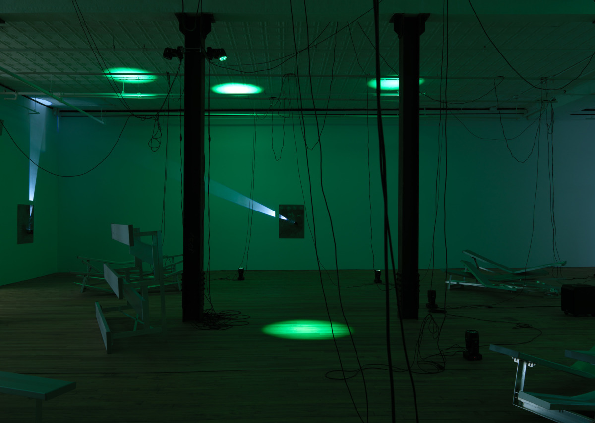a green lit room with misshapen silver bleachers arranged around the room and with many wires dangling from the ceiling