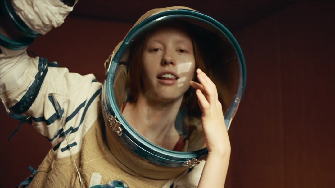 image of a white person wearing a white and blue astronaut suit and transparent glass helmet, they grin slyly at the camera and their hand caresses their helmet