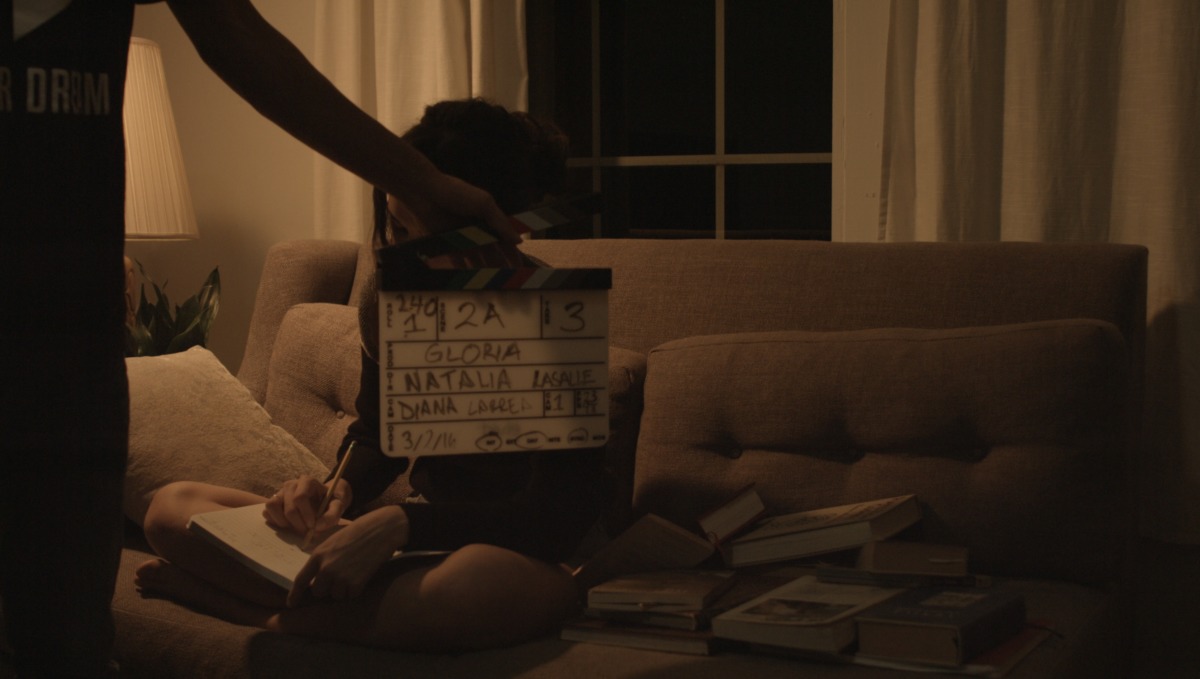 a woman sits on a couch in a dimly lit room on a beige couch. there are books next to her and she is writing in a journal. a man holds a clapperboard in front of her face.