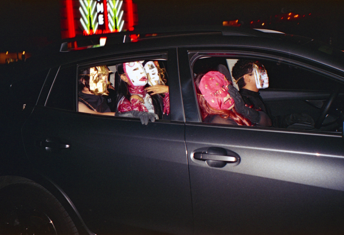 five kabuki masked people in a silver car wearing gloves drive thru a parking lot at night. 