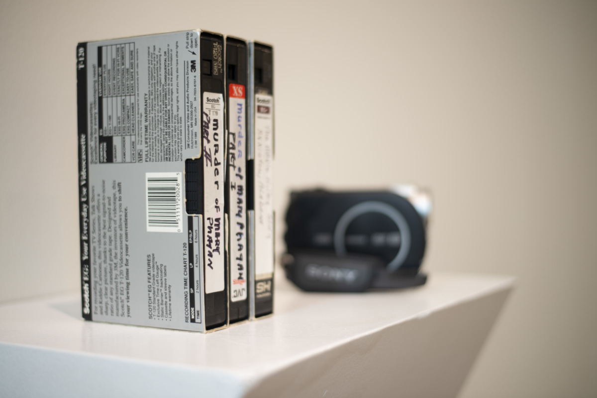on a white podium against a white wall are three stacked VHS tapes of home made movies. the labels on the VHS tapes say the murder of Mary phatah parts 1-3. In the background there is a handheld camcorder.