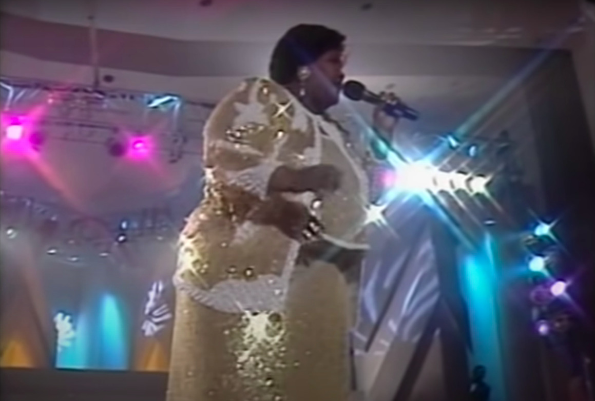 a larger-bodied Black woman wears a glittering gold and white church suit that sparkles in the spotlight, she stands on stage with a microphone in her left hand, blue and pink production lights shine behind her