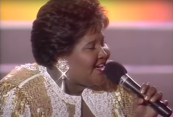 a larger-bodied Black woman wears a gold and white suit bedazzled in sparkles, that catch the light as she moves across the stage, at this moment the light catches the large pearlescent earring in her ear as she tilts the microphone toward her red lips
