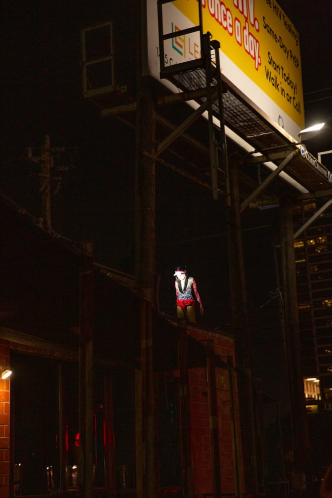 a person in a kabuki mask stands underneath a billboard at night, with light shining on their mask. 
