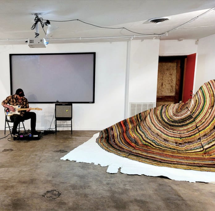 a person sits and performs a soundcheck alongside a large painting work on white canvas