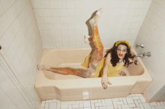 film photograph of a drag queen wearing a yellow outfit with colorful tights inside of a white bathroom in a light pink tub