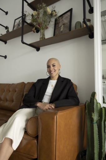 a lighter skinned Black woman with a shaved head dyed blonde. she sits on a brown leather couch wearing a white shirt, black blazer, and cream colored pants