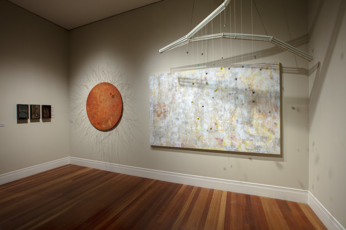 three white walls and a golden brown hard wood floor. on the center wall hangs a large orange disk with spry threads like hairs. there is a large white, yellow and gray painting to the right of it. hanging from the ceiling in front of the white and gray painting is a forked piece of metal, with dangling strings with bottle caps attached, like a mobile.  