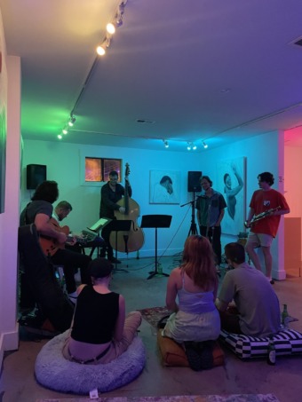 interior of a white gallery with multicolored ambient lights, a crowd surrounds a group of musicians playing jazz music in the dimly lit space