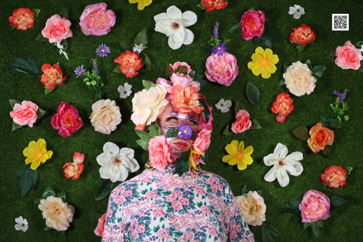 a person wearing flowers covering their face and a floral bodysuit standing in front of a grassy background with bright colored flowers pinned to the greenery
