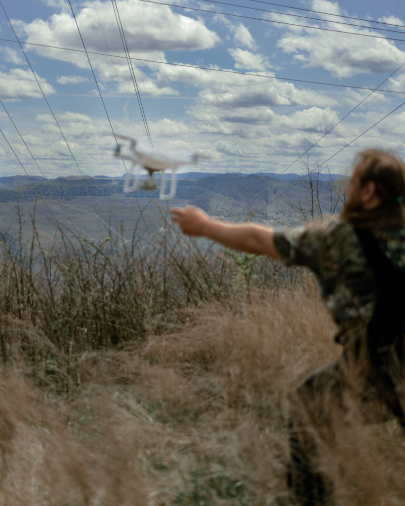 a man in a field in camp overalls releases a white aerial drone into the sky.