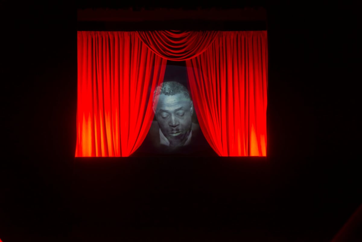 a black and white image of a black man looking down superimposed on an image of red curtains parting. 