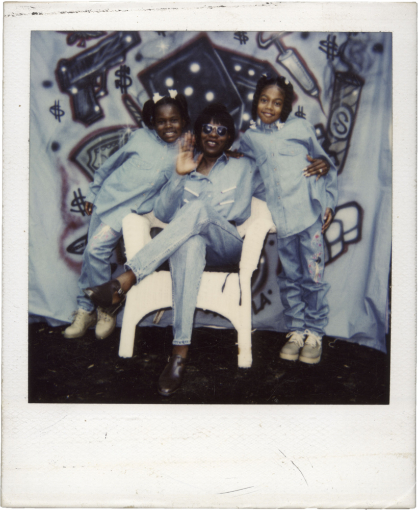 scanned polaroid of three black women, one adult and two girls to her left and right, face and pose for the camera. they wear denim suits and the adult is sitting on a white chair in front of a graffiti backdrop