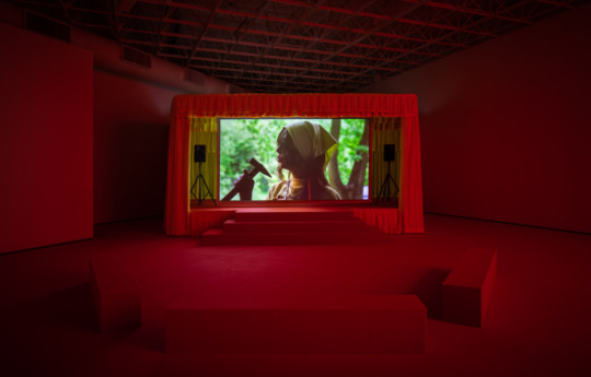 Installation view of red lit room with screen in the center, a girl holding a hammer stands projected