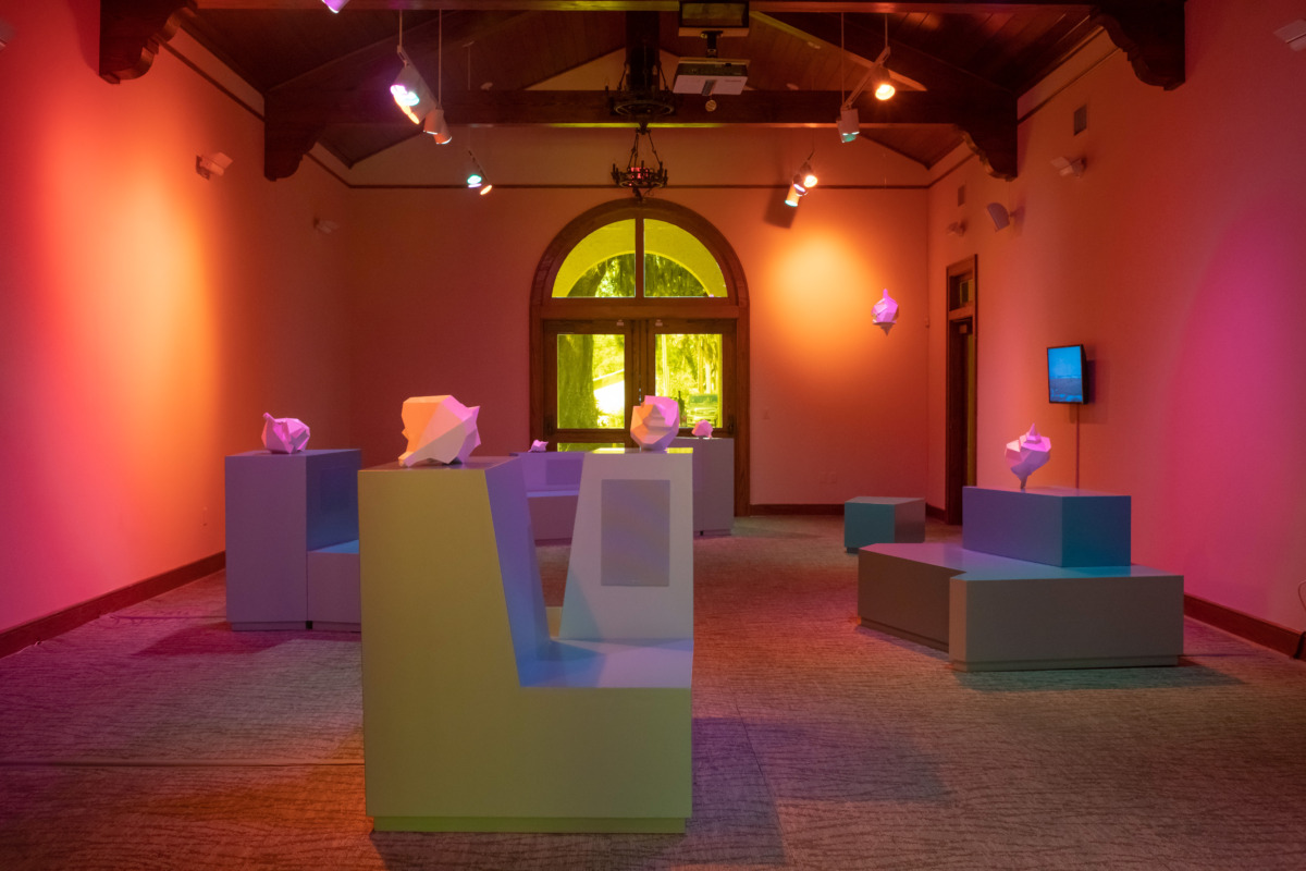 Installation view of the exhibition Dreams of Unknown Islands including grey bench sculpture with 3D printed shells, monitors, and orange/pink walls. 