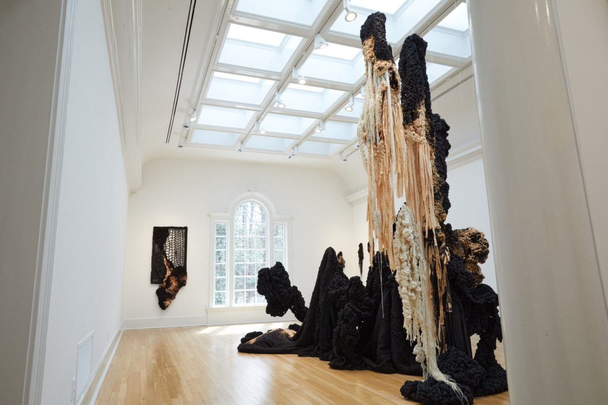 a white-walled, light-filled gallery is filled with sweeping textiles that reach from floor to ceiling, the installations are a charcoal black with white knots and tassles the color of bones