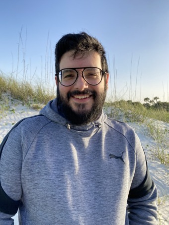 A headshot of Christopher, who is standing on a beach. He has thick black hair and a full beard that covers his light beige skin. He wears large circular glasses and a large smile on his face