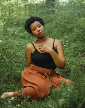 a photo of Destiny sitting in a wild field of green, Destiny has a short afro and medium brown skin, wears a black camisole and loose pants the color of red clay, the expression on Destiny's face is dreamy