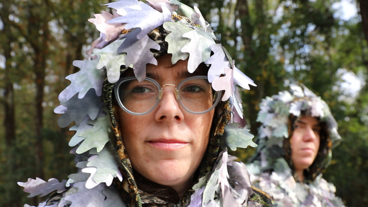 A figure wearing glasses faces the camera. An additional figure is obscured in the background. Both are wearing hoods covered in artifical flowers. 