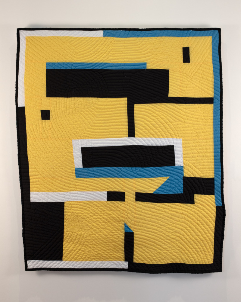 A geometric quilt comprised primarily of blocks of yellow and black with smaller blocks of white and blue. 