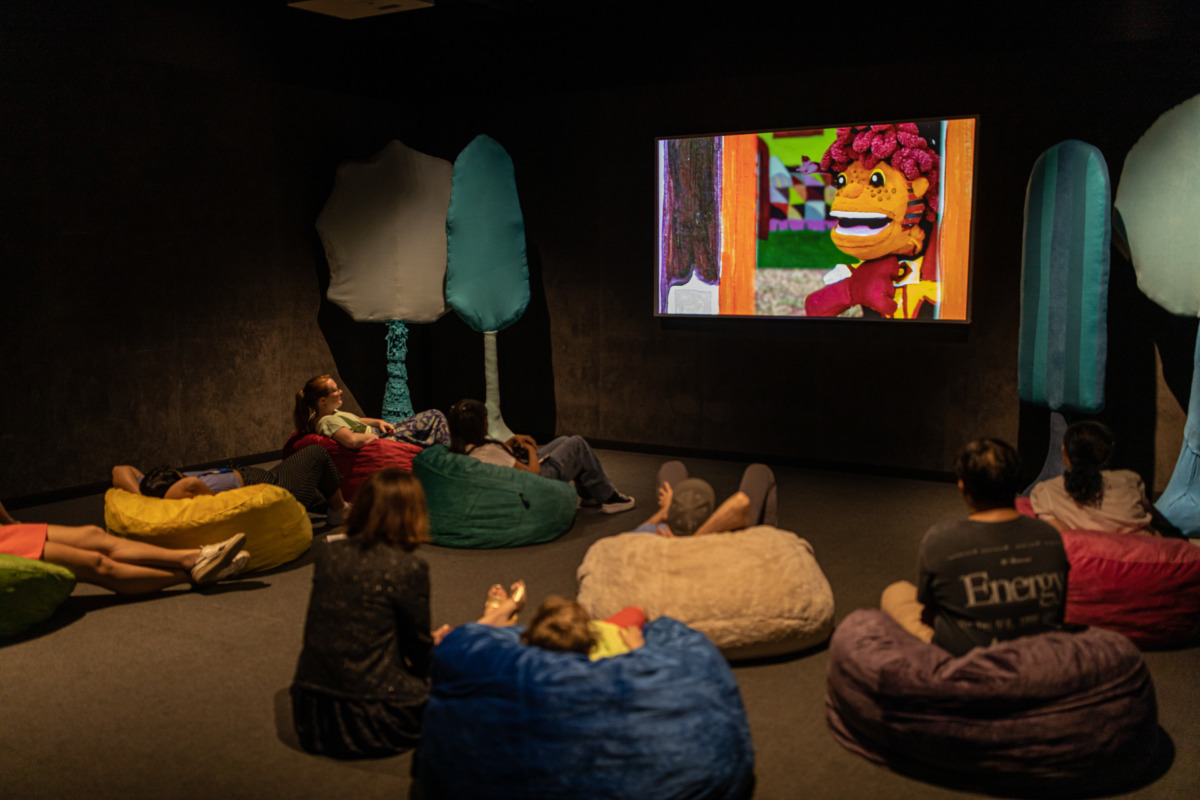 A large screen with a plush character talking on it. People sitting in beanbag chairs facing the screen.
