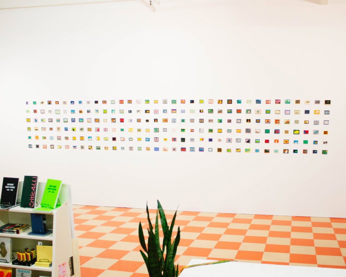 white wall of small symmetrical square artworks above floor of orange and white squares with snake plant and half of bookshelf in foreground
