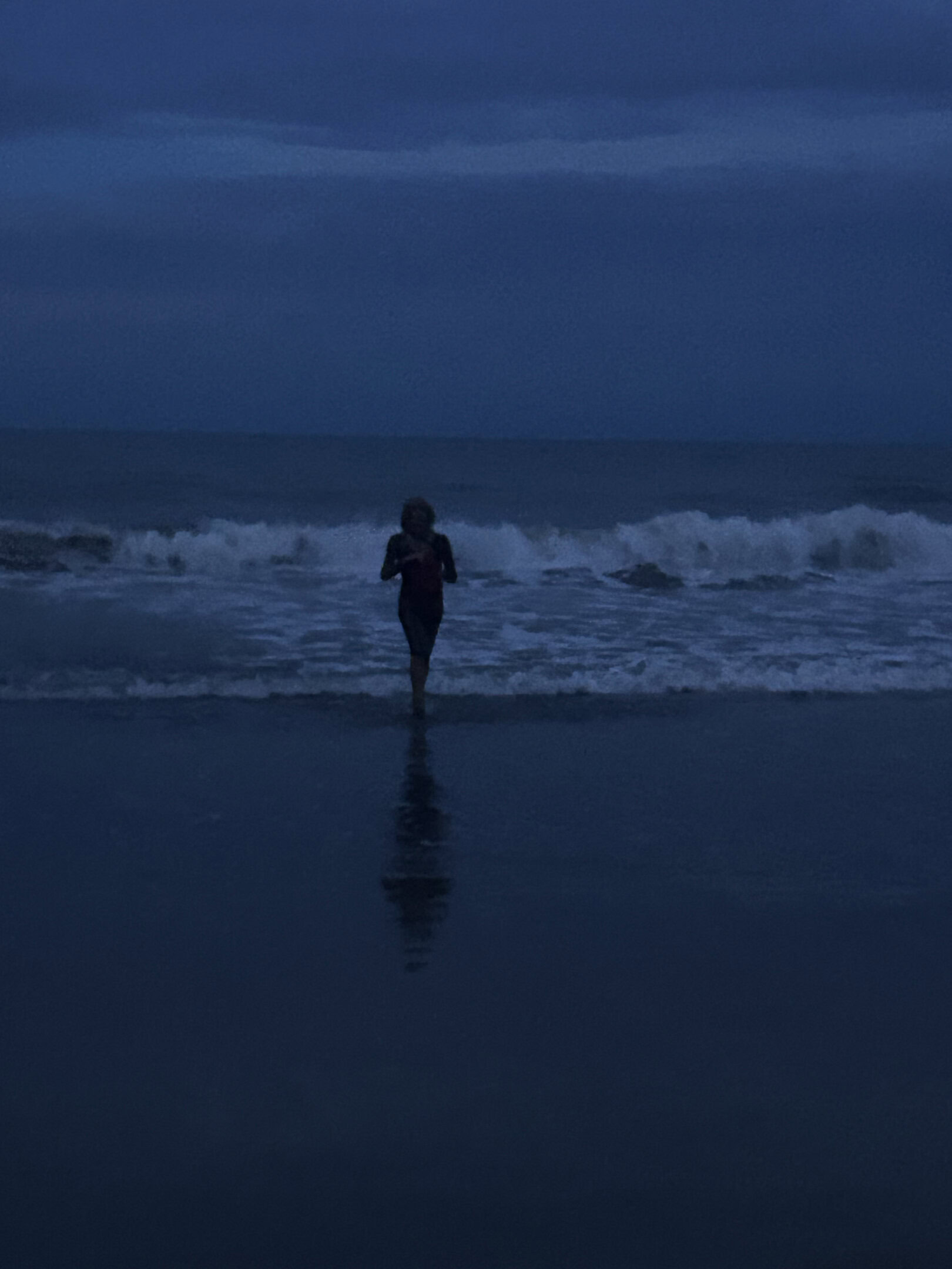 A woman walks toward the camera: the crashing blue waves of the ocean at twilight just behind her.